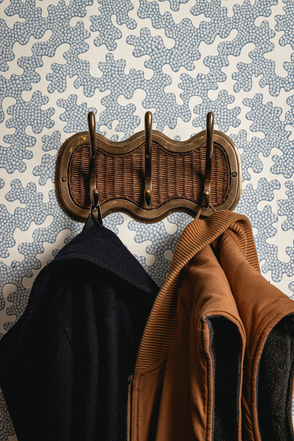 Wicker coat rack with two jackets against a patterned wall.